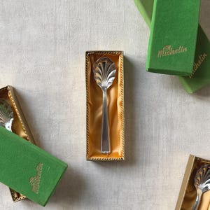Puede incluir: Una cuchara de servir plateada con un cuenco decorativo en forma de concha, presentada en una caja forrada de oro. También se ven varias cajas verdes con la palabra "Michelin".