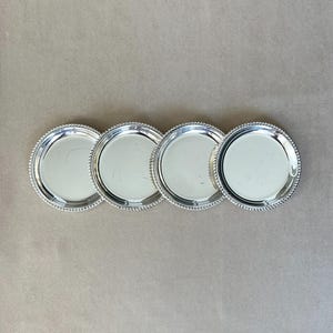 Könnte beinhalten: Vier silberfarbene, runde Untersetzer mit einem Perlenrand sind in einer Reihe angeordnet. Die Untersetzer haben eine reflektierende Oberfläche und sind vor einem neutralen, hellbraunen Hintergrund platziert. Diese Untersetzer eignen sich für die Inneneinrichtung.
