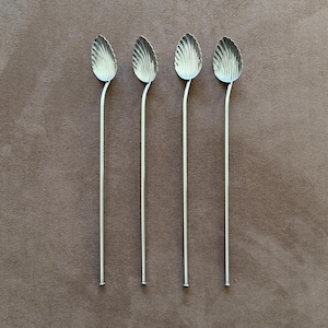 May include: Four silver spoons with leaf-shaped bowls and long, slender handles. The spoons are arranged in a row against a brown background. The spoons are likely for serving or stirring beverages.