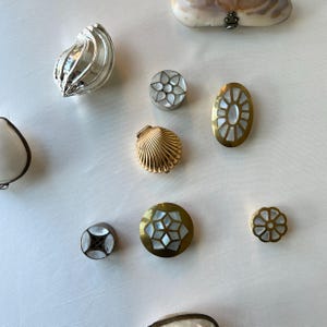 May include: An assortment of decorative shell-themed objects, including a silver clam shell, a gold scallop shell, and various round and oval pieces with mother-of-pearl inlays. The pieces are in gold, silver, and white.