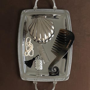 Könnte beinhalten: Ein silbernes rechteckiges Tablett mit Griffen enthält einen muschelförmigen Behälter, eine Perlenkette, eine Parfümflasche mit schwarzer Quaste, eine rechteckige Parfümflasche und einen dunklen Kamm. Das Tablett hat einen Perlenrand.