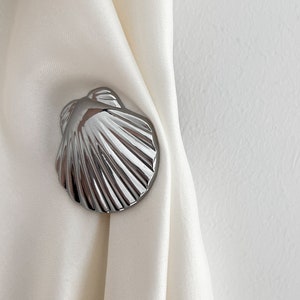 Può includere: Un fermaglio per tende a forma di conchiglia argentata. Il fermaglio è a forma di conchiglia di capesante con una finitura liscia e lucida.