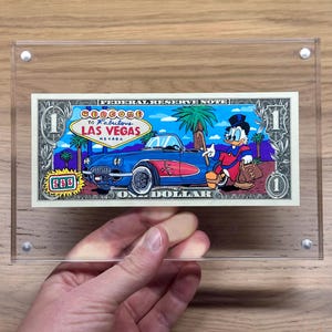 Könnte beinhalten: Ein gerahmter Ein-Dollar-Schein mit einem blauen Cabrio, Palmen und dem Text "Welcome to Fabulous Las Vegas Nevada". Die Rechnung enthält auch eine Zeichentrickfigur, die eine Geldbörse hält.