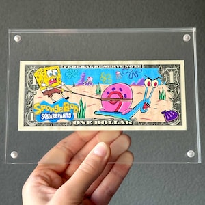 Può includere: Un biglietto da un dollaro incorniciato con una scena di cartoni animati disegnata a mano di SpongeBob che tira Gary la lumaca. L'opera d'arte è in una cornice di acrilico trasparente, con le parole "SpongeBob SquarePants" e "One Dollar".