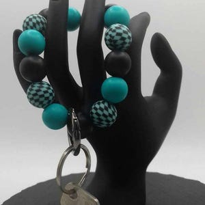May include: A beaded keychain wristlet in black, teal, and checkered patterns. The wristlet is attached to a silver key ring with a key. The wristlet is displayed on a black hand-shaped stand.