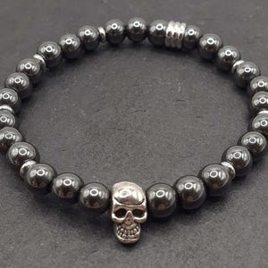 Puede incluir: Una pulsera de cuentas grises oscuras con un dije de calavera plateada. La pulsera está hecha de cuentas esféricas y ensartada en una banda elástica. El dije de calavera tiene cuencas de ojos detalladas, una cavidad nasal y dientes.