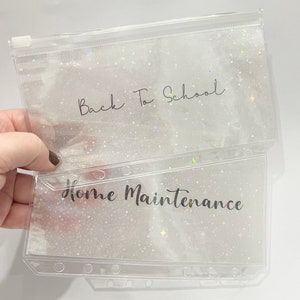 May include: Two clear plastic zippered pouches with a glittery background. The top pouch has the text "Back To School" written on it. The bottom pouch has the text "Home Maintenance" written on it.