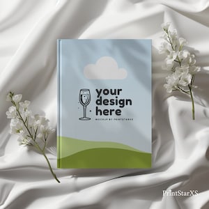 May include: A white book mockup with a blue sky and green hills background. The book has a glass of champagne icon and the text "your design here" with the text "MOCKUP BY PRINTSTARXS" below.