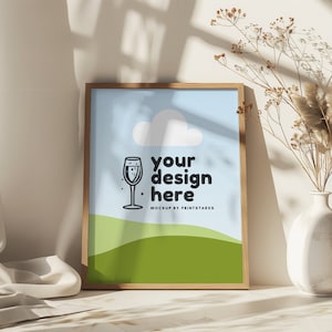 May include: A framed mockup with a light blue background, a white cloud, a green hill, and a black and white illustration of a glass of champagne. The text "your design here" is in black and the text "MOCKUP BY PRINTSTARXS" is in grey.