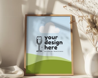 May include: A framed mockup with a light blue background, a white cloud, a green hill, and a black and white illustration of a glass of champagne. The text "your design here" is in black and the text "MOCKUP BY PRINTSTARXS" is in gray.