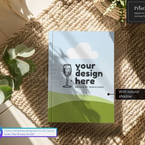K&ouml;nnte beinhalten: Ein Mockup eines Buchcovers mit hellblauem Hintergrund und einer gr&uuml;nen und wei&szlig;en Landschaft. Der Text "your design here" ist in Schwarz auf dem Cover geschrieben. Das Buch liegt auf einem beigefarbenen gewebten Teppich mit einem nat&uuml;rlichen Schatten.
