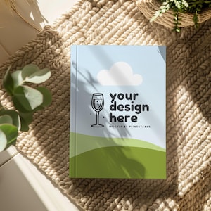 K&ouml;nnte beinhalten: Ein blaues Buch-Mockup mit einer wei&szlig;en Wolke und einer Illustration eines Weinglases. Der Text "your design here" ist auf dem Buch gedruckt. Das Buch liegt auf einem beigefarbenen Strickteppich.