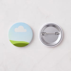 May include: Two round pin badges. One badge features a blue sky with a white cloud and green hills. The other badge shows the silver pin back. The badges are approximately 3.8 cm in diameter.