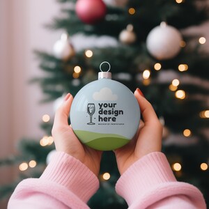 以下が含まれることがあります： 白い雲と黒い文字「your design here」が書かれた青いクリスマスオーナメント。オーナメントは人の手に持たれている。