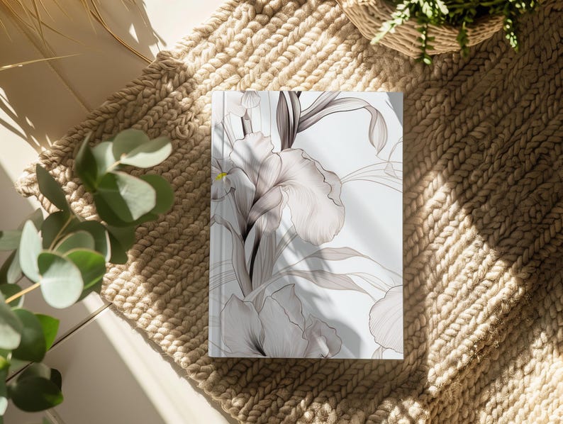 K&ouml;nnte beinhalten: Ein wei&szlig;es Journal mit einem floralen Design mit zarten grauen und wei&szlig;en Blumen. Das Journal liegt auf einem gewebten beigefarbenen Teppich.