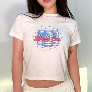 Puede incluir: Una camiseta blanca recortada con un gráfico de tres latas de refresco con el diseño de la bandera estadounidense y el texto "Ameri Can". La camiseta la lleva puesta una persona con un bolso de mano plateado.