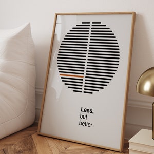 Op de afbeelding: Een ingelijste print met een geometrisch ontwerp in zwart en wit in de vorm van een cirkel. De cirkel heeft een dunne oranje lijn in het midden. De tekst "Less, but better" is onder de cirkel gedrukt.