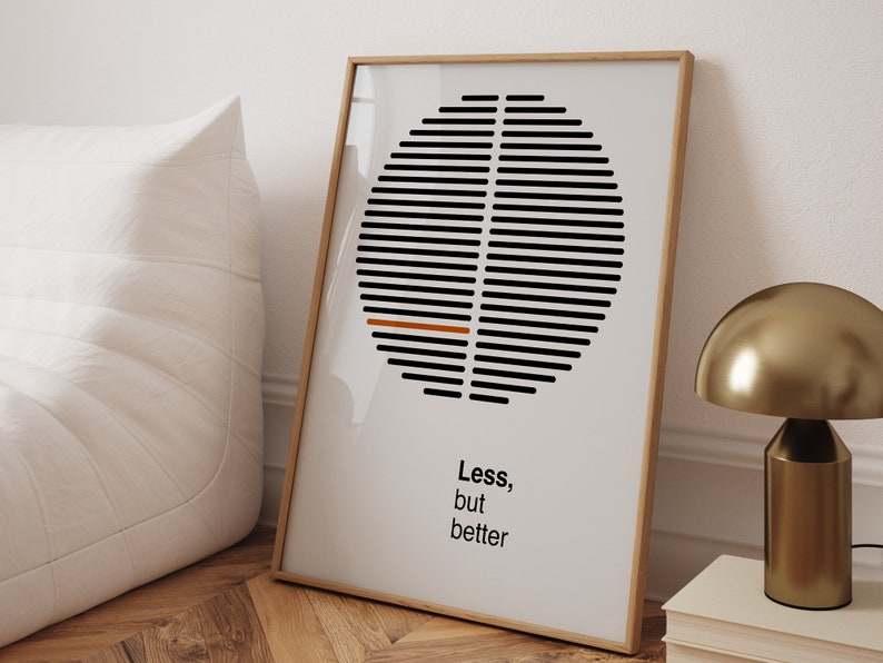 Op de afbeelding: Een ingelijste print met een geometrisch ontwerp in zwart en wit in de vorm van een cirkel. De cirkel heeft een dunne oranje lijn in het midden. De tekst "Less, but better" is onder de cirkel gedrukt.