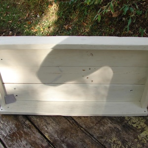 May include: A rectangular, off-white wooden shelf with a shallow depth. The shelf is constructed from horizontal planks and has a simple, clean design. The wood has a slightly distressed appearance.