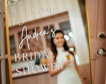 Może przedstawiać: Lustro w złotej ramie z napisem "Welcome to Andrea's Bridal Shower" napisanym białymi literami. Kobieta w białej sukience odbija się w lustrze.