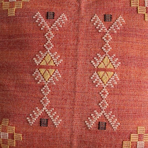 May include: Close-up of a woven textile with a repeating pattern. The background is a reddish-orange color. The pattern consists of white, yellow, and black geometric shapes. The design is symmetrical and appears to be embroidered.