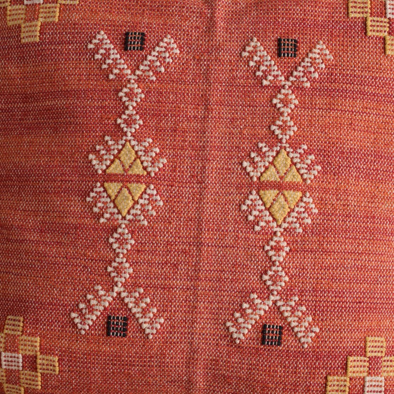 May include: Close-up of a woven textile with a repeating pattern. The background is a reddish-orange color. The pattern consists of white, yellow, and black geometric shapes. The design is symmetrical and appears to be embroidered.