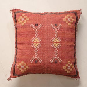 May include: A square, coral-colored decorative pillow with a geometric pattern. The pillow features a central design of vertical, stylized floral motifs in white and yellow, framed by a border with tassels at each corner. The pillow is made of woven fabric.