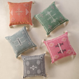 May include: Five square decorative throw pillows in shades of coral, green, blue, pink, and gray. Each pillow features a geometric embroidered design in white and cream, with a tassel at each corner. The pillows are arranged on a neutral surface.
