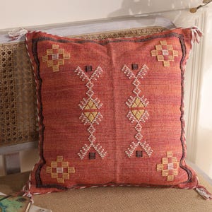 May include: A square, rust-colored decorative pillow with a geometric pattern in white, yellow, and black. The pillow has tassels on each corner and is placed on a light-colored wooden bench.