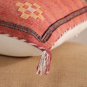 May include: Close-up of a decorative pillow with a textured, woven orange and red fabric. The pillow features a geometric pattern in yellow and black, with a white backing and a tassel detail. The pillow is resting on a tan surface.