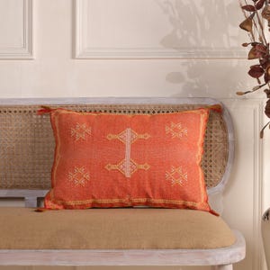 May include: An orange rectangular cushion with a geometric pattern. The cushion is placed on a light brown bench with a woven back. The cushion has a textured appearance and is trimmed with a gold border. The cushion measures approximately 40cm x 60cm.