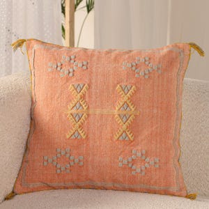 Può includere: Cuscino decorativo quadrato color pesca con motivi geometrici ricamati in giallo e grigio. Il cuscino ha un bordo giallo e nappe su ogni angolo. Il cuscino è su un divano bianco.
