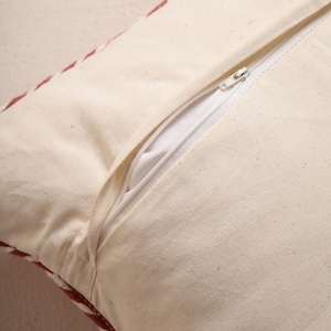 May include: Close-up of a cream-colored pillow cover with a white zipper partially open, revealing a white interior. The pillow has a red and white patterned border.