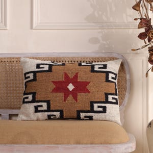 Puede incluir: Un cojín rectangular de textura tejida con un motivo central de estrella roja de ocho puntas. El diseño incluye patrones geométricos escalonados en blanco y negro sobre un fondo color canela, creando una apariencia simétrica y multicolor.