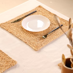 Puede incluir: Un mantel individual de yute tejido con un plato blanco, un tenedor y un cuchillo. El mantel individual es de forma cuadrada con una textura trenzada. El tenedor y el cuchillo tienen mangos negros. El plato es blanco y redondo.