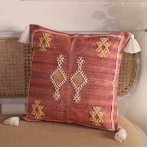Op de afbeelding: Een vierkant decoratief kussen met een rood-oranje geweven stof. Het kussen heeft witte en gele geometrische borduursels en witte kwastjes op elke hoek. Het kussen ligt op een bruin oppervlak.