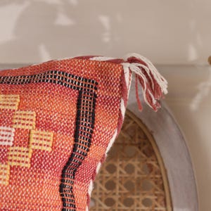 May include: Close-up of a woven pillow with a geometric pattern in shades of orange, red, black, and yellow. The pillow has a white fringe along the edge. The pillow is part of a home decor collection.