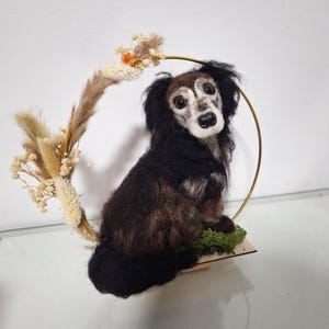 Könnte beinhalten: Eine Nadelfilz-Hundeskulptur mit schwarzem und braunem Fell, die auf einem Holzsockel mit Moos sitzt. Die Skulptur ist von einem goldenen Reifen umgeben, der mit getrockneten Blumen verziert ist.