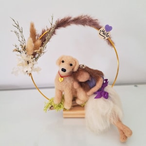 Könnte beinhalten: Handgefertigte Filzhundefigur mit dem Aussehen eines Golden Retrievers, mit rotem Halsband und goldenem herzförmigen Anhänger. Der Hund befindet sich in einem goldenen Reifen, der mit Trockenblumen und einem Holzschild mit der Aufschrift "Meilo" verziert ist.