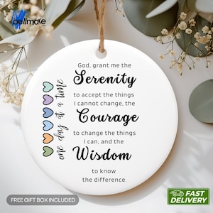Op de afbeelding: Een rond wit keramisch ornament hangend aan een bruin touwtje. De tekst luidt: God, grant me the Serenity to accept the things I cannot change, the Courage to change the things I can, and the Wisdom to know the difference. One day at a time.