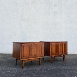 May include: Two mid-century modern wooden cabinets. Each cabinet has a rectangular shape with four doors featuring a geometric design. The cabinets are supported by four tapered legs and a curved base. The wood has a warm, brown tone.