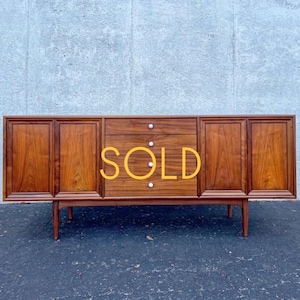 May include: A long wooden sideboard featuring a rich brown wood grain finish, four cabinet doors with recessed panels, and a central column of four drawers with small white circular knobs. The word SOLD is printed in yellow across the center.