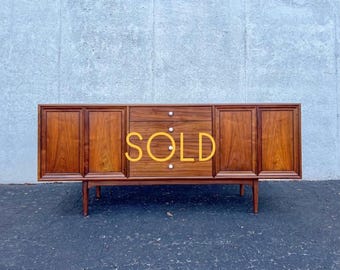 May include: A long wooden sideboard featuring a rich brown wood grain finish, four cabinet doors with recessed panels, and a central column of four drawers with small white circular knobs. The word SOLD is printed in yellow across the center.