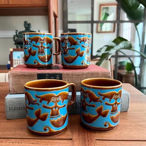 May include: Four ceramic mugs with a mermaid design. The mugs have a brown rim and handle, with a turquoise blue body featuring brown mermaid illustrations. The mugs are stacked on a wooden surface.