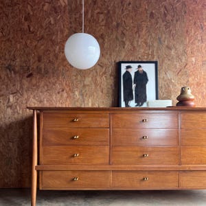 May include: A mid-century modern wooden dresser with multiple drawers and brass knobs. A white globe pendant light hangs above. A framed black and white print and decorative items sit on top of the dresser.
