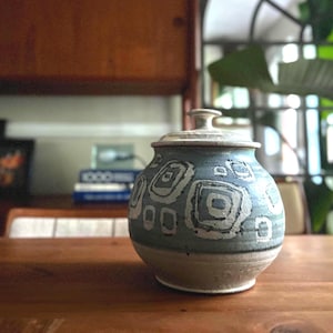 May include: A round ceramic jar with a lid, featuring a light blue and cream speckled glaze. The exterior is decorated with a repeating pattern of dark gray and white hand-painted squares and rectangles arranged in a geometric, abstract design.