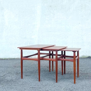 May include: A set of three nesting tables crafted from medium-toned wood. Each table features a rectangular top with rounded edges and four tapered legs connected by curved support stretchers, allowing the smaller tables to slide underneath.