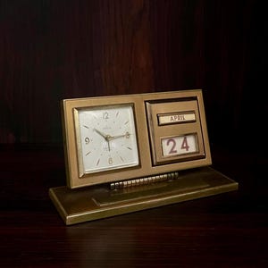 May include: A vintage gold-toned desk clock with a square clock face and a calendar display. The clock face has black numerals and hands. The calendar shows "April 24". The clock is set on a rectangular base.