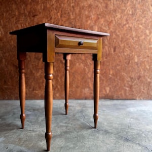 May include: A small, wooden side table with a single drawer and four turned legs. The table is stained in a warm, medium-brown color, and the drawer has a small, round knob. The table's dimensions are approximately 24 inches high, 18 inches wide, and 12 inches deep.