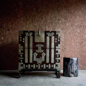 May include: A dark wood chest with brass hardware and accents, including vertical bars, diamond shapes, and a central lock. A small, dark, cylindrical stool sits to the right. The chest is on a black base.
